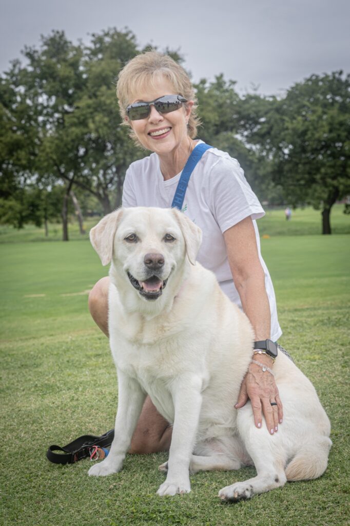 A woman in sunglasses kneeling on the grass beside a smiling yellow Lab