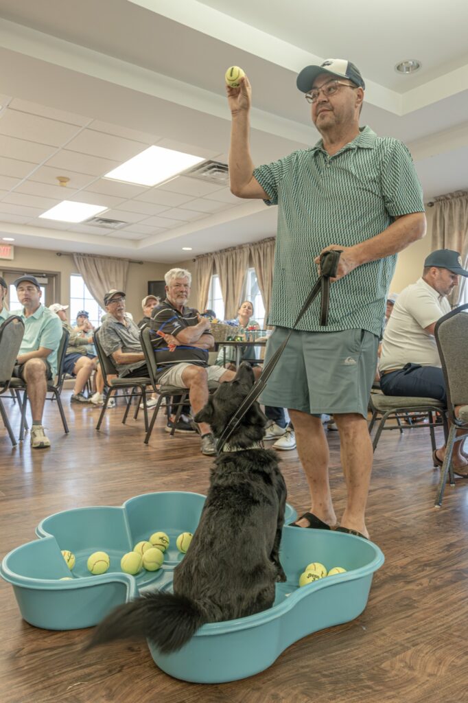 A man holds a tennis ball above a black dog standing in a bone-shaped pool of tennis balls while seated guests watch