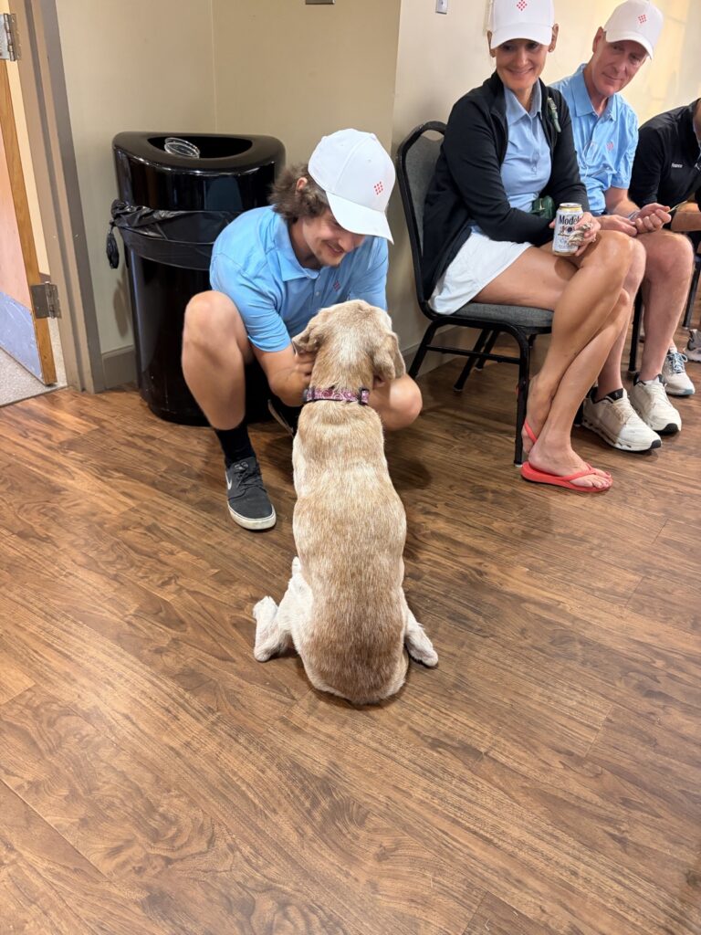 A golfer in a blue shirt kneels to pet a yellow Lab indoors while other players sit nearby